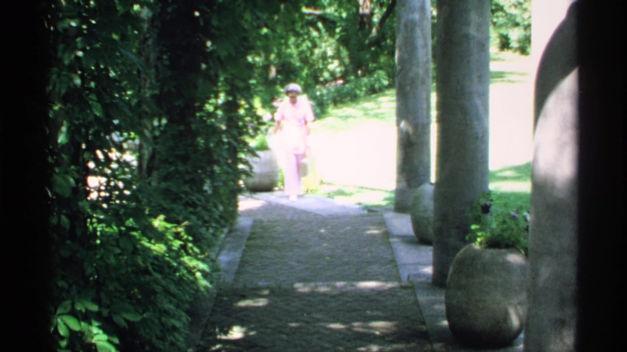 In 1970 Minnesota home-movie footage, a woman in pink appears on a stone path framed by foliage and columns.