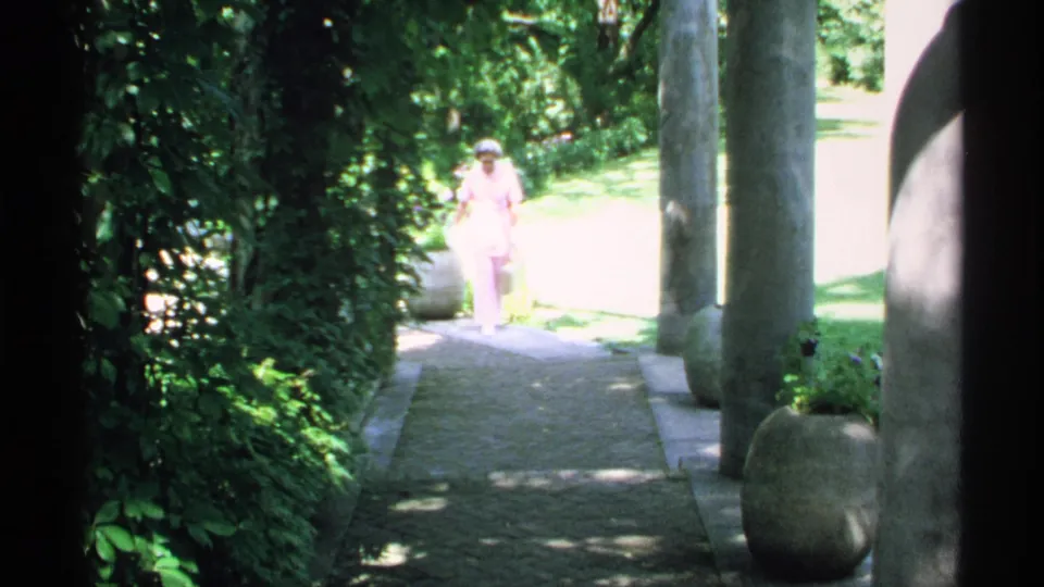 In 1970 Minnesota home-movie footage, a woman in pink appears on a stone path framed by foliage and columns.