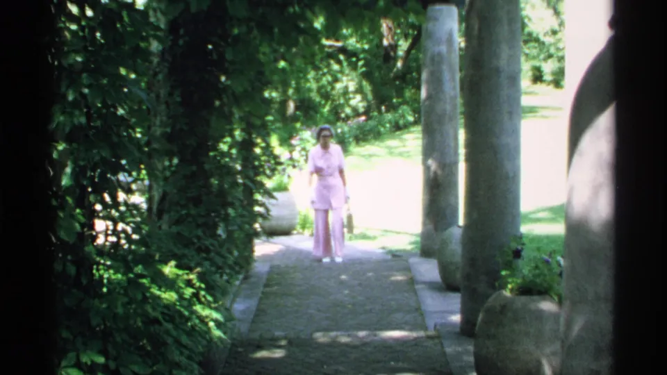 A woman in a pink outfit walks along a sun-dappled garden path in Minnesota 1970 8mm footage.