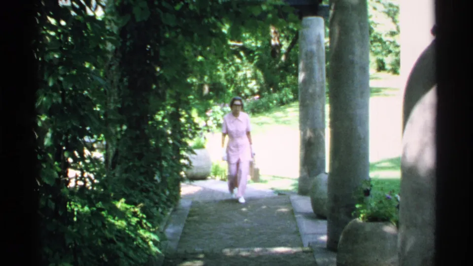 The woman walks closer to the camera on a shaded stone walkway, framed by garden plants and columns.