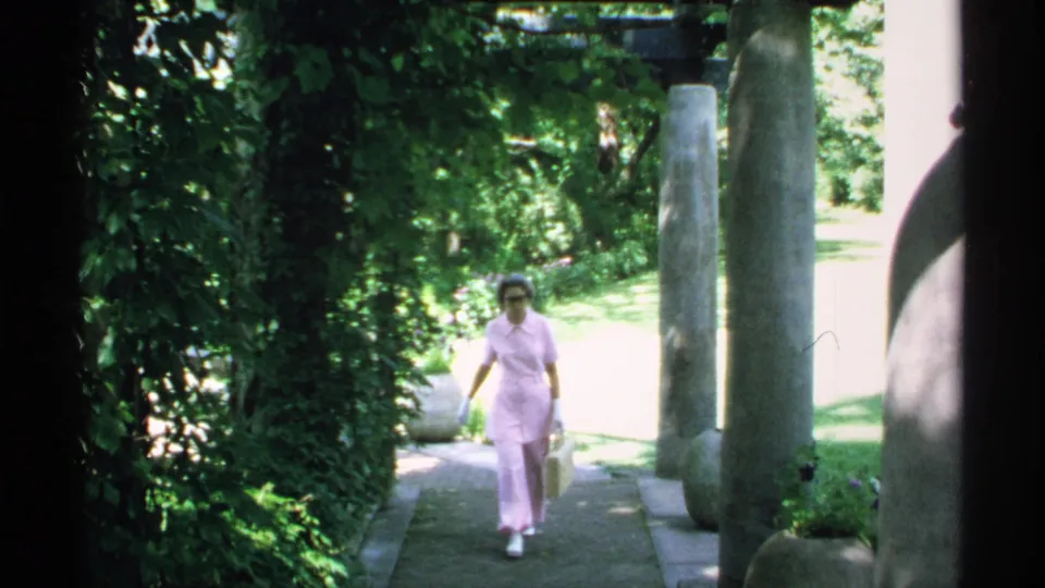 Near the end of the 1970 Minnesota clip, the woman carries a pale handbag while walking through the green garden walkway.