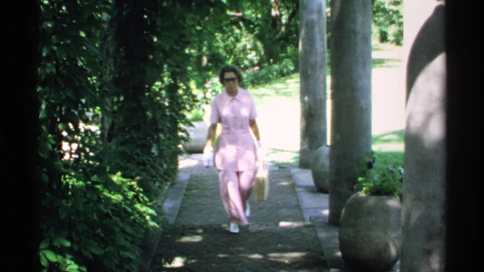 A woman in a pink suit and white shoes walks toward the camera on a landscaped Minnesota path in 1970 home-movie footage.