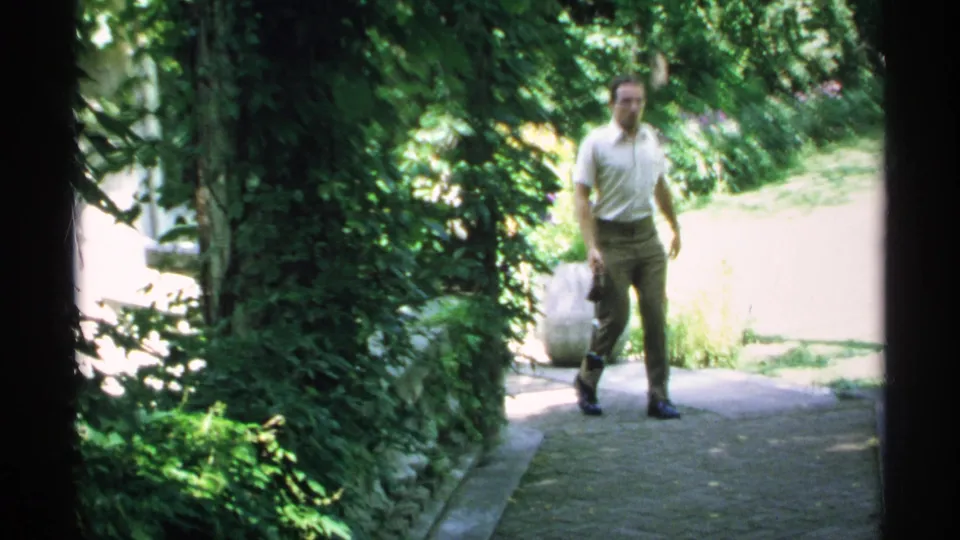 A man in a light shirt and brown trousers walks toward the camera along the shaded garden path in 1970 Minnesota footage.