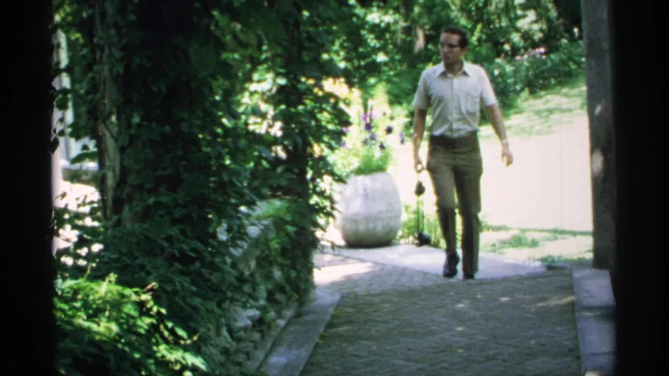 The man approaches on the same landscaped path, framed by greenery, a stone pillar, and a large planter in 1970 Minnesota home-movie footage.