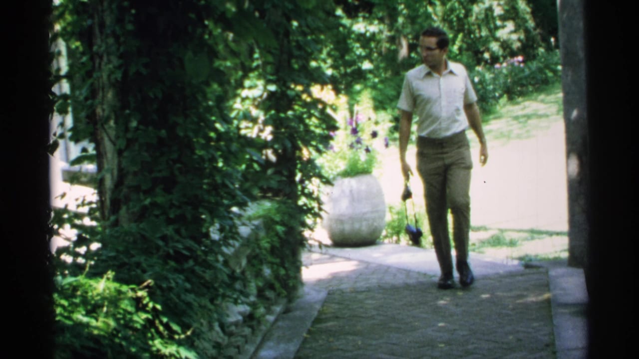 In Minnesota around 1970, a man in a light shirt walks down a sun-dappled garden path with greenery and a stone planter nearby.