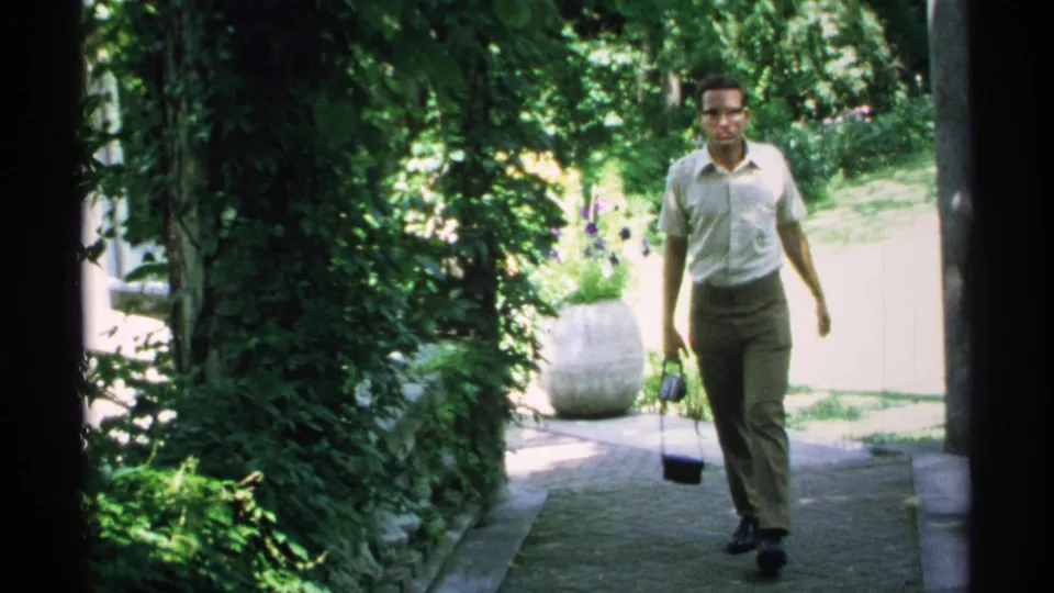 The man continues toward the camera on a Minnesota garden path, framed by leafy plants, a stone planter, and bright background light.