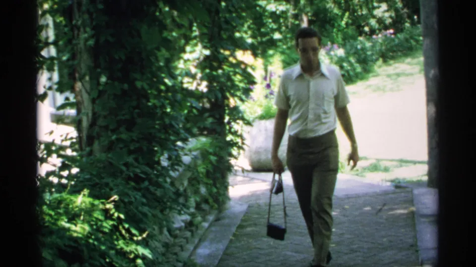 A closer view shows the man walking past dense greenery and an ivy-covered wall, carrying a small bag in one hand.