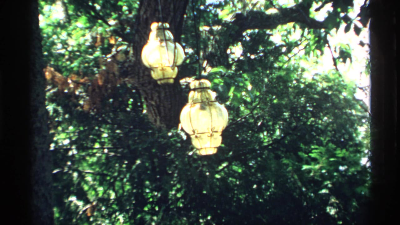 1970 Minnesota 8mm frame showing two ornate hanging lanterns suspended from a tree against dense green backyard foliage.
