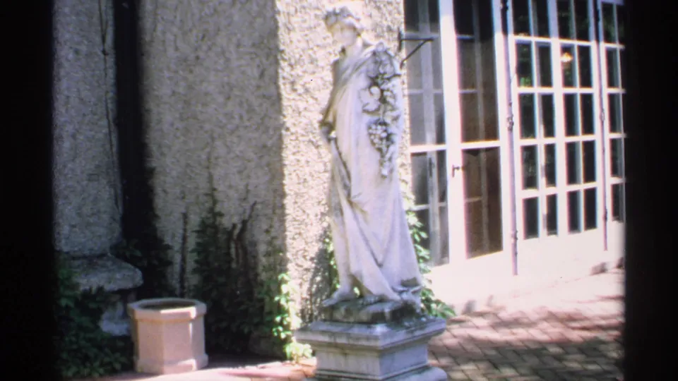 1970 Minnesota 8mm frame showing a classical robed statue on a pedestal near stucco walls, French doors, and brick patio paving.