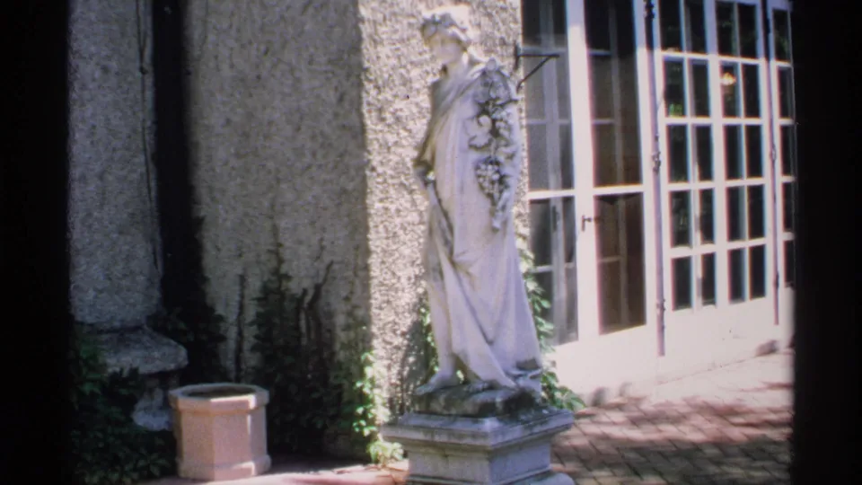 1970 Minnesota home-movie frame of a robed patio statue beside French doors, textured stucco, brick paving, and garden growth.
