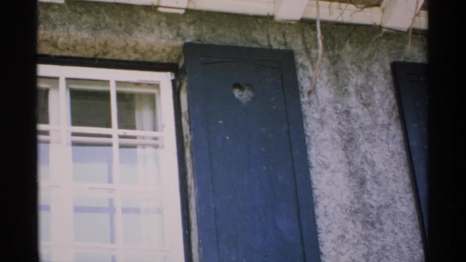 1970 Minnesota exterior detail with a pale multi-pane window and dark shutter marked by a heart-shaped cutout.