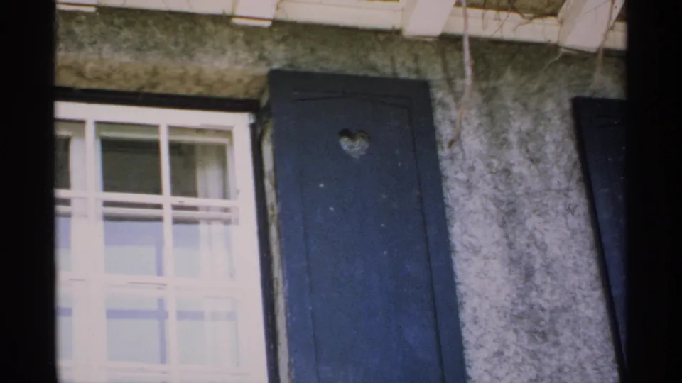 1970 Minnesota 8mm frame holding on a house exterior with window, curtain, and dark shutter with heart cutout.