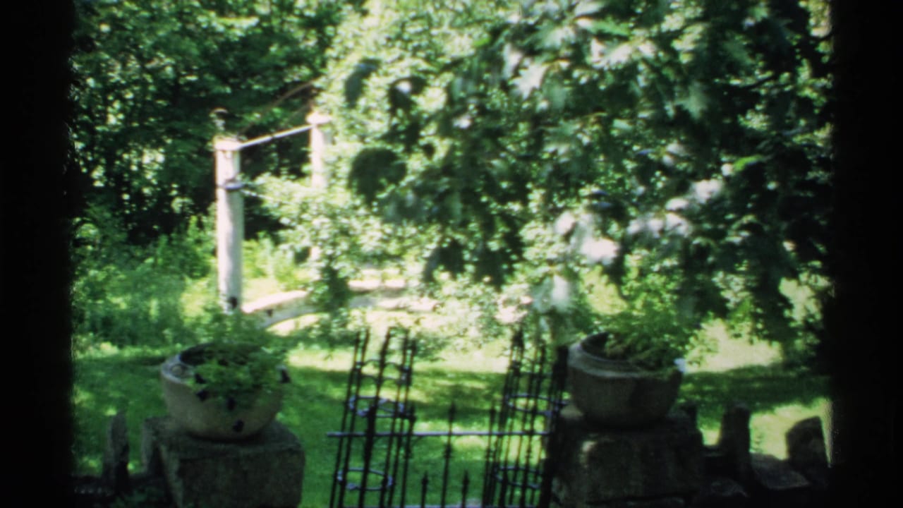 Leafy garden seen through a dark frame with stone planters, lawn, fencing, and a white arched bridge.