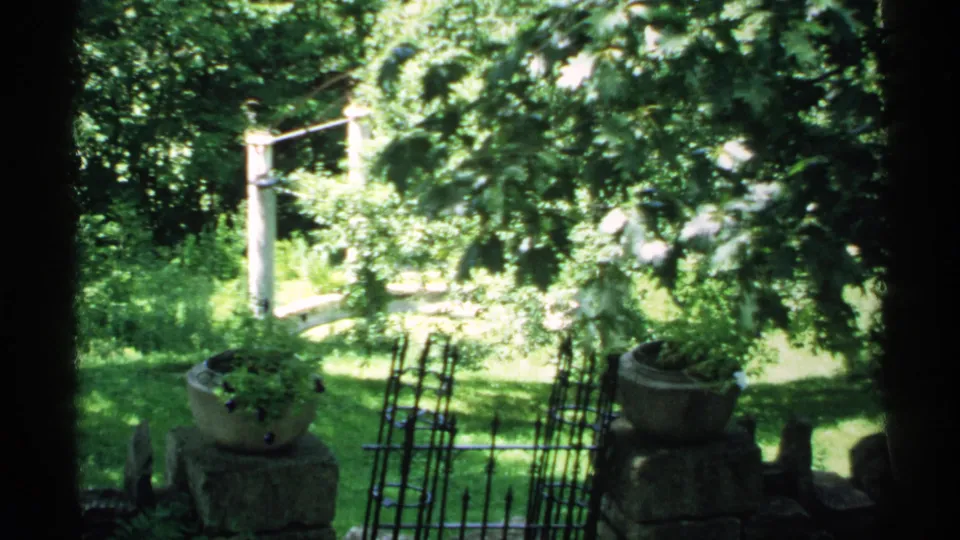 1970 Minnesota garden scene in 8mm footage, framed by dark vertical edges, showing a stone planter, iron fencing, green lawn, trees, and a white bridge.