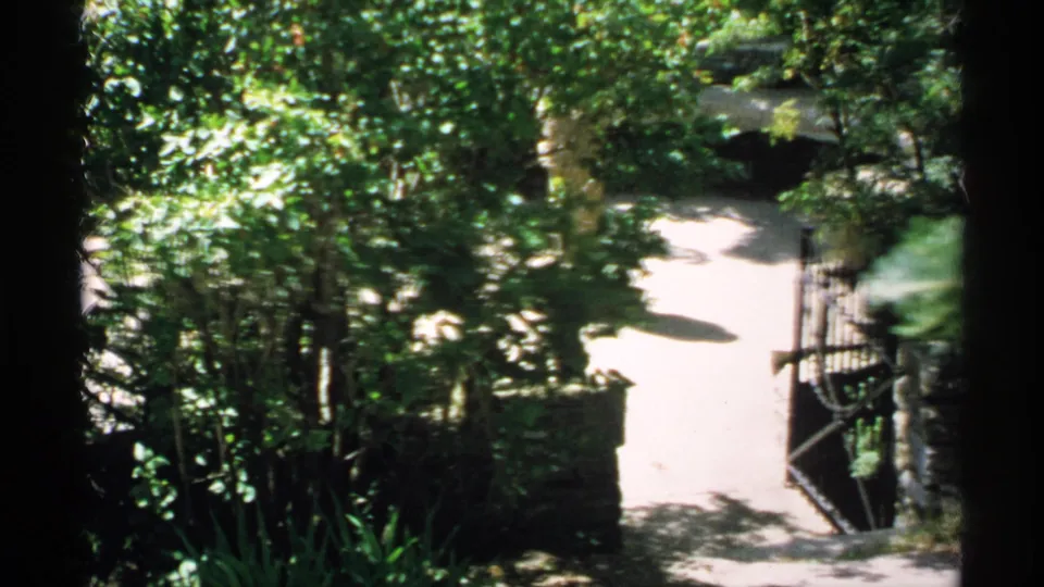 1970 Minnesota 8mm view looking down toward a bright garden path, open wrought iron gate, stone posts, shrubs, and heavy tree shade.