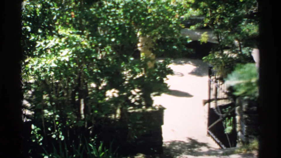 1970 Minnesota home-movie frame of a landscaped garden path with an open iron gate, stone posts, dense shrubs, and dappled tree shade.