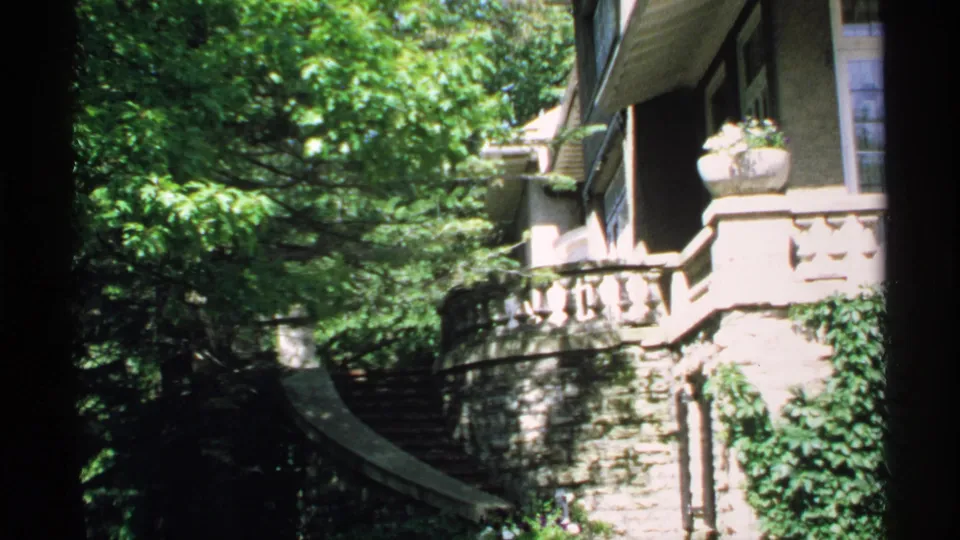 1970 Minnesota 8mm frame showing a curved stone staircase, ivy-covered masonry, balcony railing, and house exterior in sun and shade.