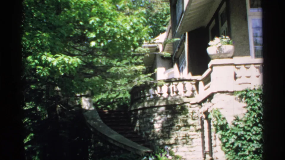 1970 Minnesota 8mm frame of a stone house exterior with ivy, curved steps, balcony railing, trees, and a hanging or mounted planter.