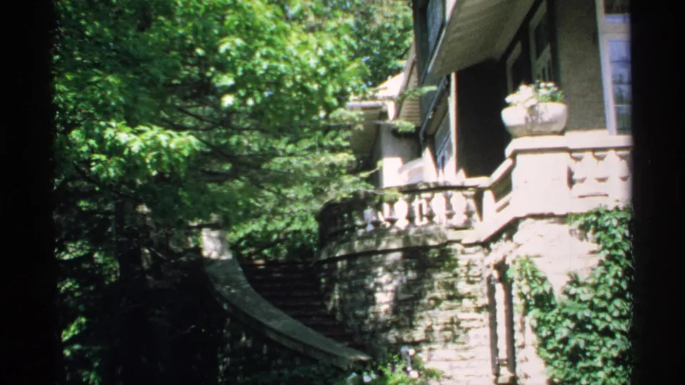 1970 Minnesota 8mm frame showing a leafy exterior view of a stone mansion with curved steps, balustrades, and white-trimmed windows.