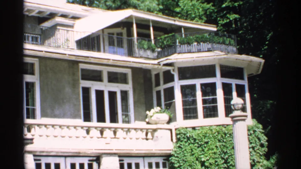 1970 Minnesota 8mm frame showing the mansion's upper balcony, white-trimmed bay window, porch railings, and surrounding trees.
