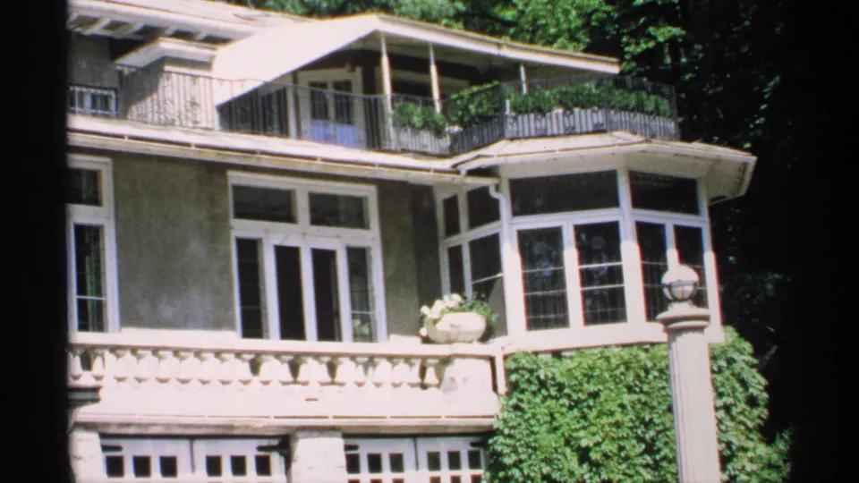 1970 Minnesota 8mm frame with a closer look at the mansion facade, balcony railings, large windows, ivy, and a decorative lamp post.