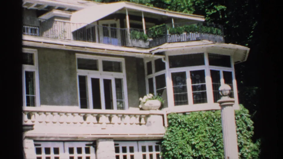 1970 Minnesota 8mm frame showing the mansion's bay window, balcony railing, stone balustrade, ivy growth, and tree-shaded exterior.