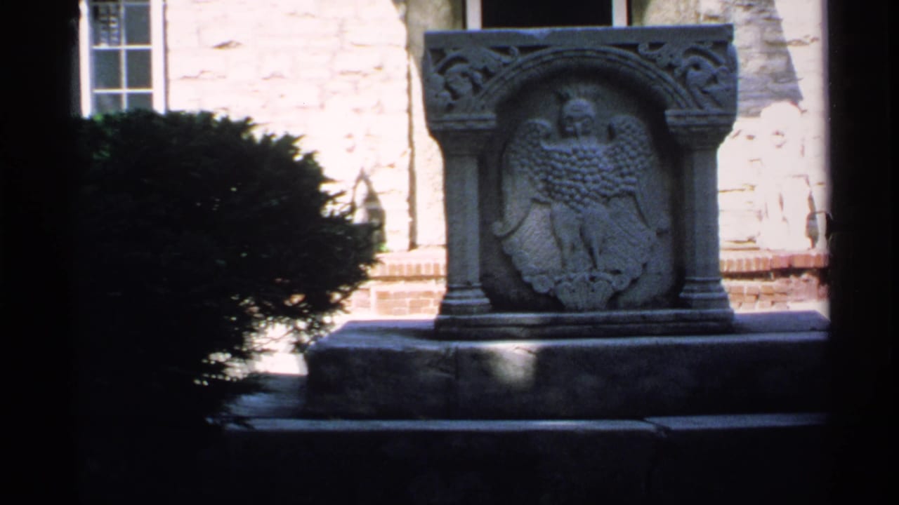 Carved stone eagle monument in front of a pale masonry wall with shrubbery at the side.