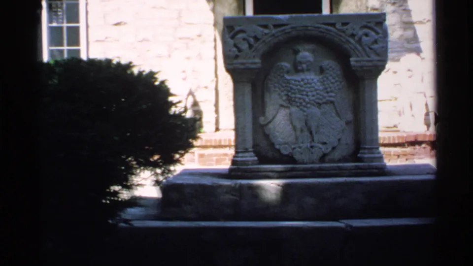 1970 Minnesota 8mm frame showing a carved stone eagle monument against a pale masonry building, partly framed by dark foreground edges and shrubs.