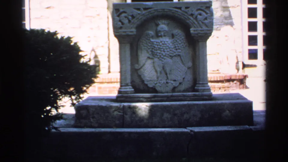 The camera holds on the carved stone eagle monument in a Minnesota home-movie shot, with shrubs and a building window visible behind it.