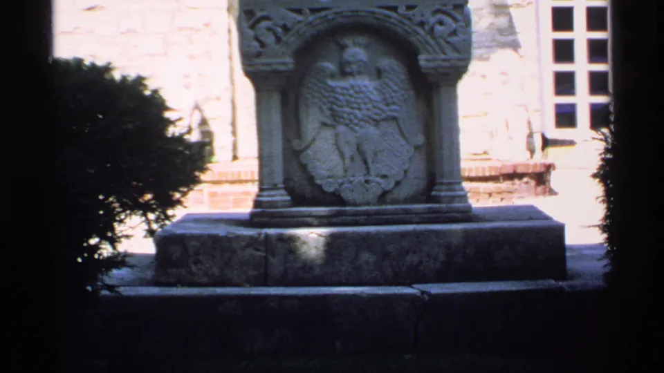 A closer 1970 Minnesota frame shows the carved eagle emblem on a stone monument, with a shrub and building window at the edges of the composition.