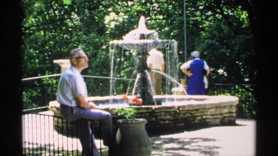 1970 Minnesota park scene with a man seated near a railing while two adults stand by a multi-tiered fountain under bright sun and trees.