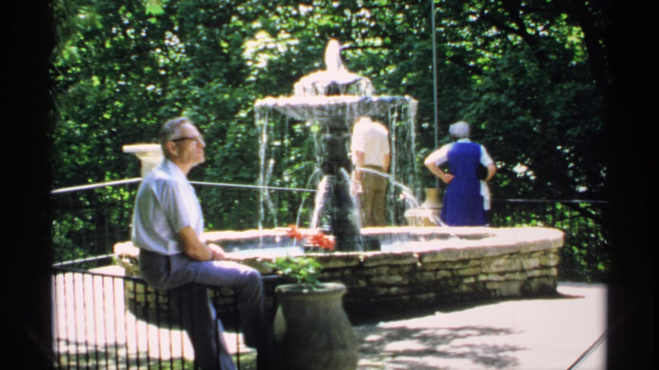 Older man sits near a stone fountain while two visitors stand nearby in a wooded park.