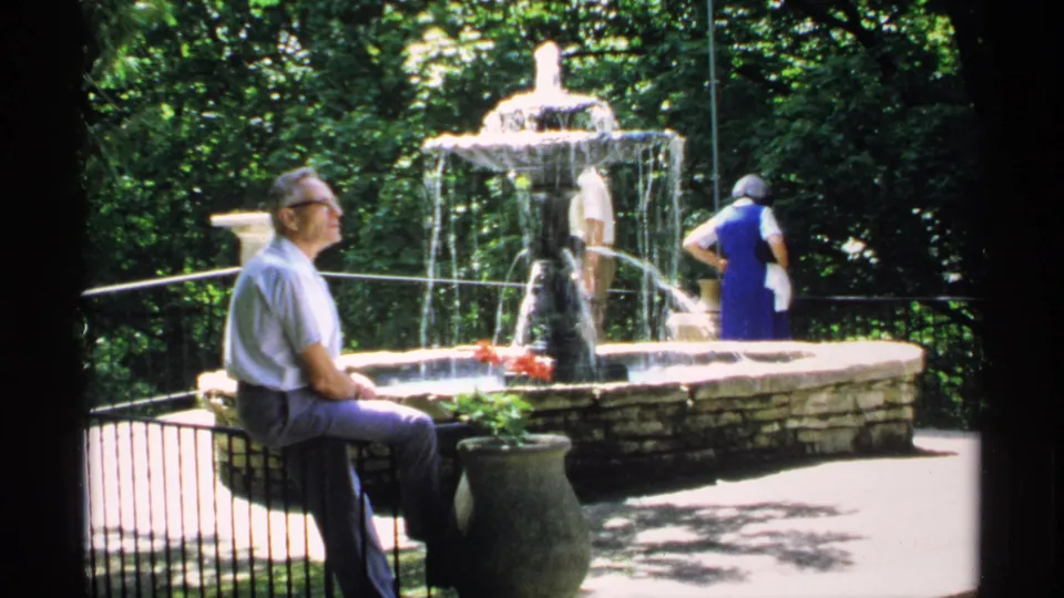 Older man in glasses sits by a sunlit stone fountain in a Minnesota park, with two visitors partly visible nearby.
