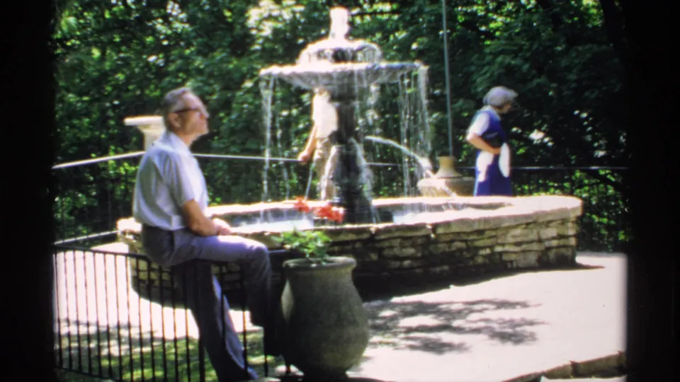 1970 Minnesota park frame showing an older man seated near a fountain while a woman in blue stands beside the stonework.