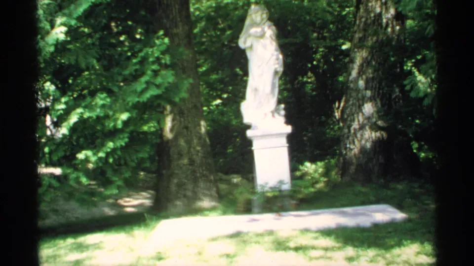 1970 Minnesota 8mm frame showing a white robed statue on a pedestal in a sunlit wooded garden.