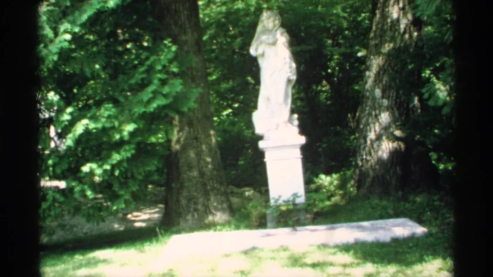 1970 Minnesota 8mm frame of a white pedestal statue set among large trees and dense summer greenery.
