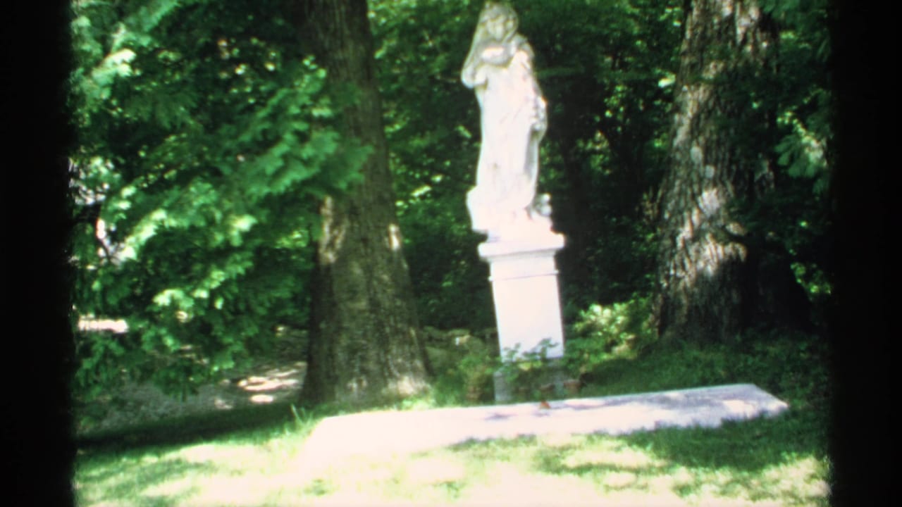 White robed statue on a pedestal among trees and grass in bright sunlight.