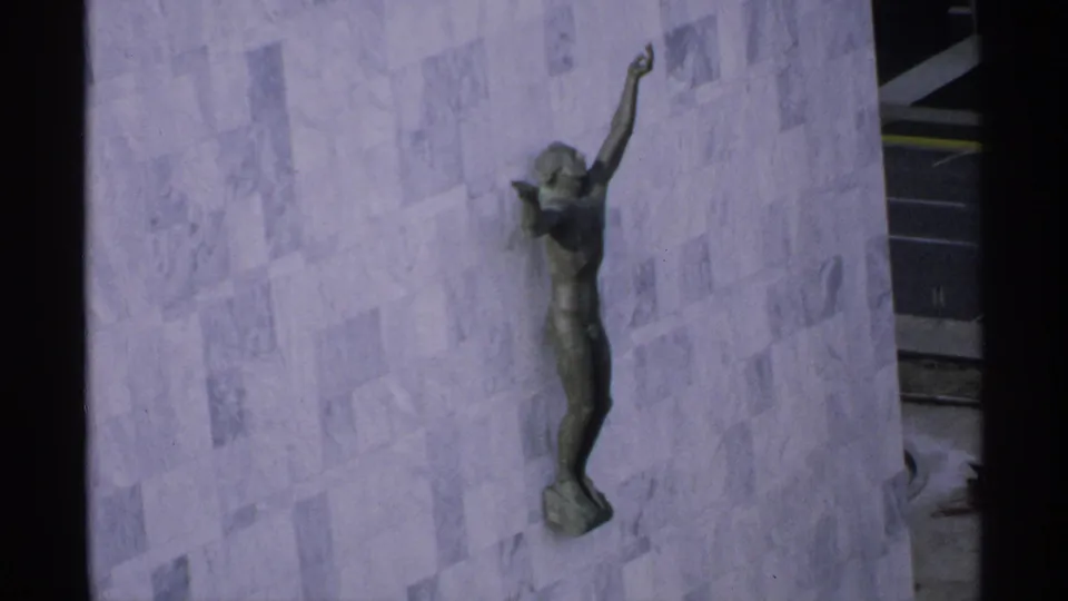 1970 Minnesota 8mm frame showing a bronze wall-mounted figure with one arm raised against a pale stone or marble surface.