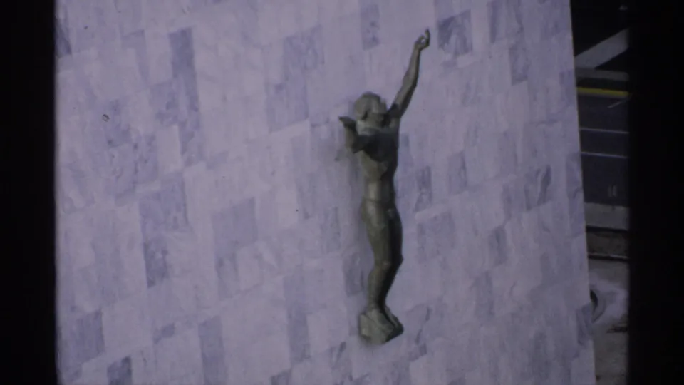 1970 Minnesota 8mm frame of a bronze figure sculpture mounted on a pale stone wall, with a dark edge of the building visible.