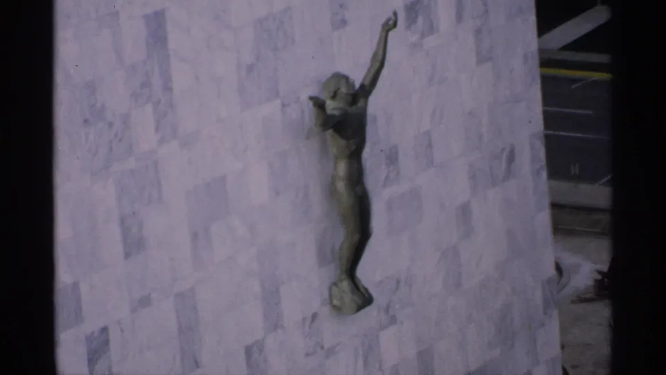1970 Minnesota home-movie frame showing a bronze figure sculpture with one raised arm on a pale stone-clad building exterior.