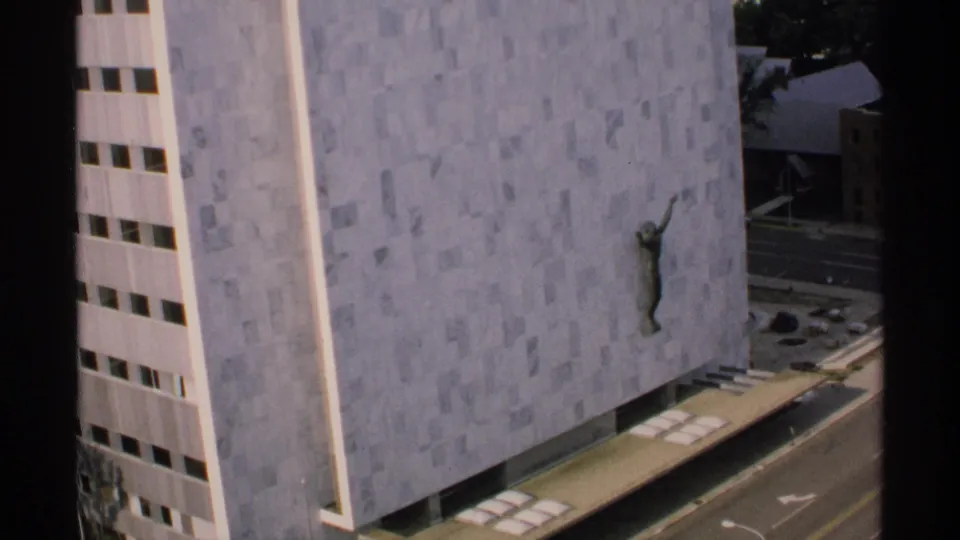 1970 Minnesota frame showing the wall sculpture on a large pale facade beside a vertical strip of windows and an urban street.