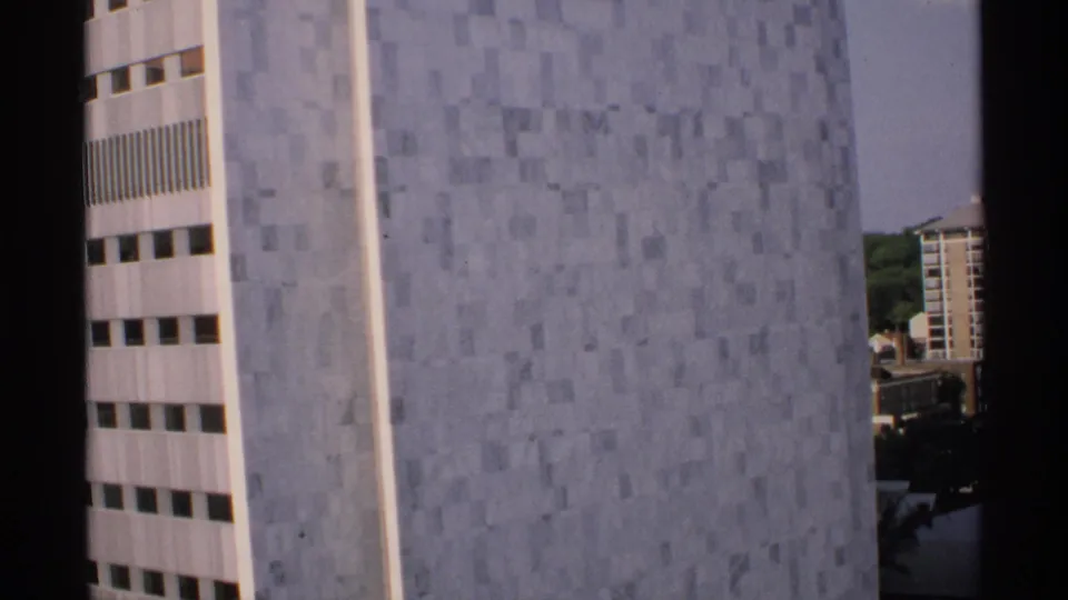 1970 Minnesota 8mm frame showing a high-rise exterior with gray square tiles, white vertical edges, and distant buildings beyond the facade.