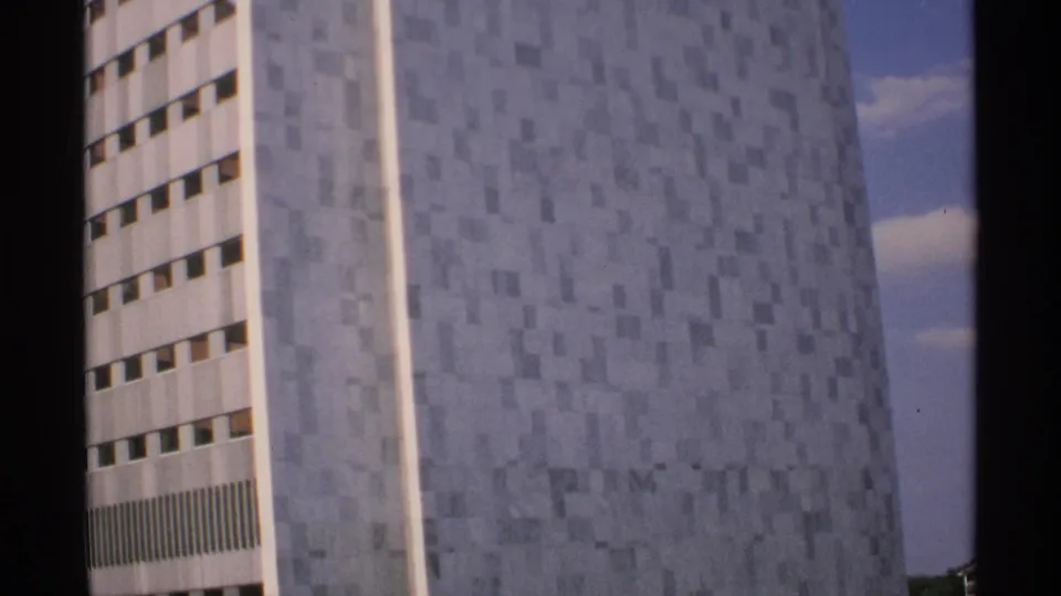 1970 Minnesota home-movie frame of a modern high-rise facade, showing patterned gray tiles, a white window bay, blue sky, and film-edge shadow.