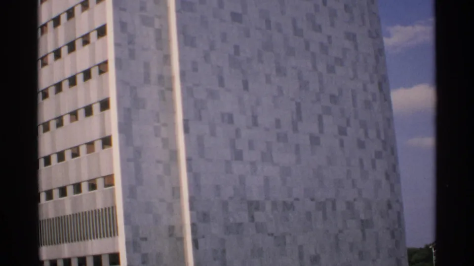 Final 1970 Minnesota 8mm frame showing the tiled skyscraper facade at a slight angle, with window rows, blue sky, clouds, and distant greenery.