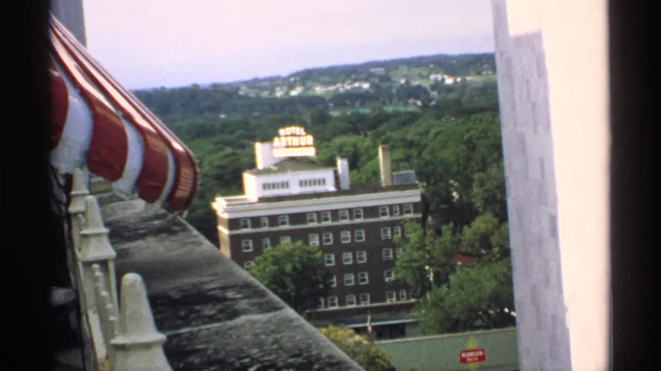 1970 Minnesota view of the Hotel Arthur sign and brick hotel facade, framed by a balcony edge, awning, and surrounding greenery.