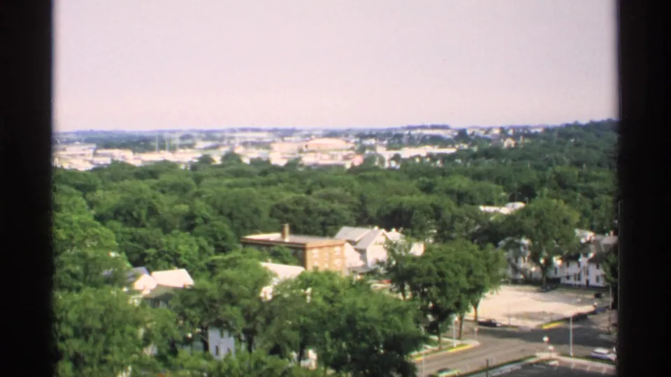 1970 Minnesota 8mm frame showing a broad high-angle view over residential houses, roads, trees, and distant low-rise development.