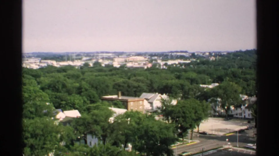 1970 Minnesota high-vantage frame looking across green residential blocks, quiet roads, and distant low-rise structures.