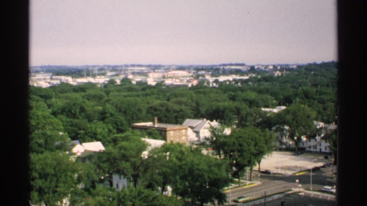 High view over a tree-filled town with houses, roads, and distant low buildings framed by dark window edges.