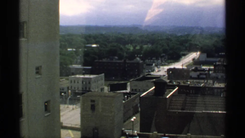1970 Minnesota high-window view with an adjacent wall, commercial rooftops, a light-toned street, dense trees, and a hazy horizon.