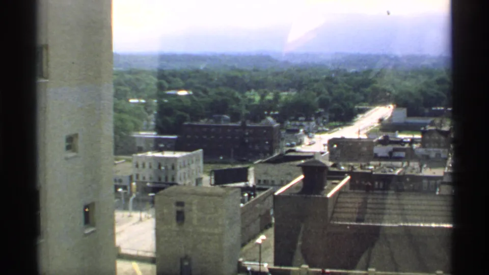1970 Minnesota 8mm frame looking from a high window over low-rise buildings, streets, treetops, and a hazy skyline.